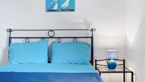 Una cama con ropa de cama azul, un cuadro de veleros en la pared y una mesita de noche con lámpara.