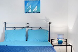 A bed with blue bedding, a painting of sailboats on the wall, and a bedside table with a lamp.