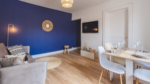 En moderne stue med en blå accentvæg, et spiseområde med et træbord og hvide stole, og et tv ophængt på væggen.