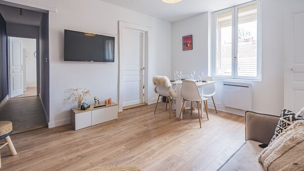 En moderne stue med et fladskærms-tv, et spiseområde med stole og et bord, og et stort vindue der giver naturligt lys.