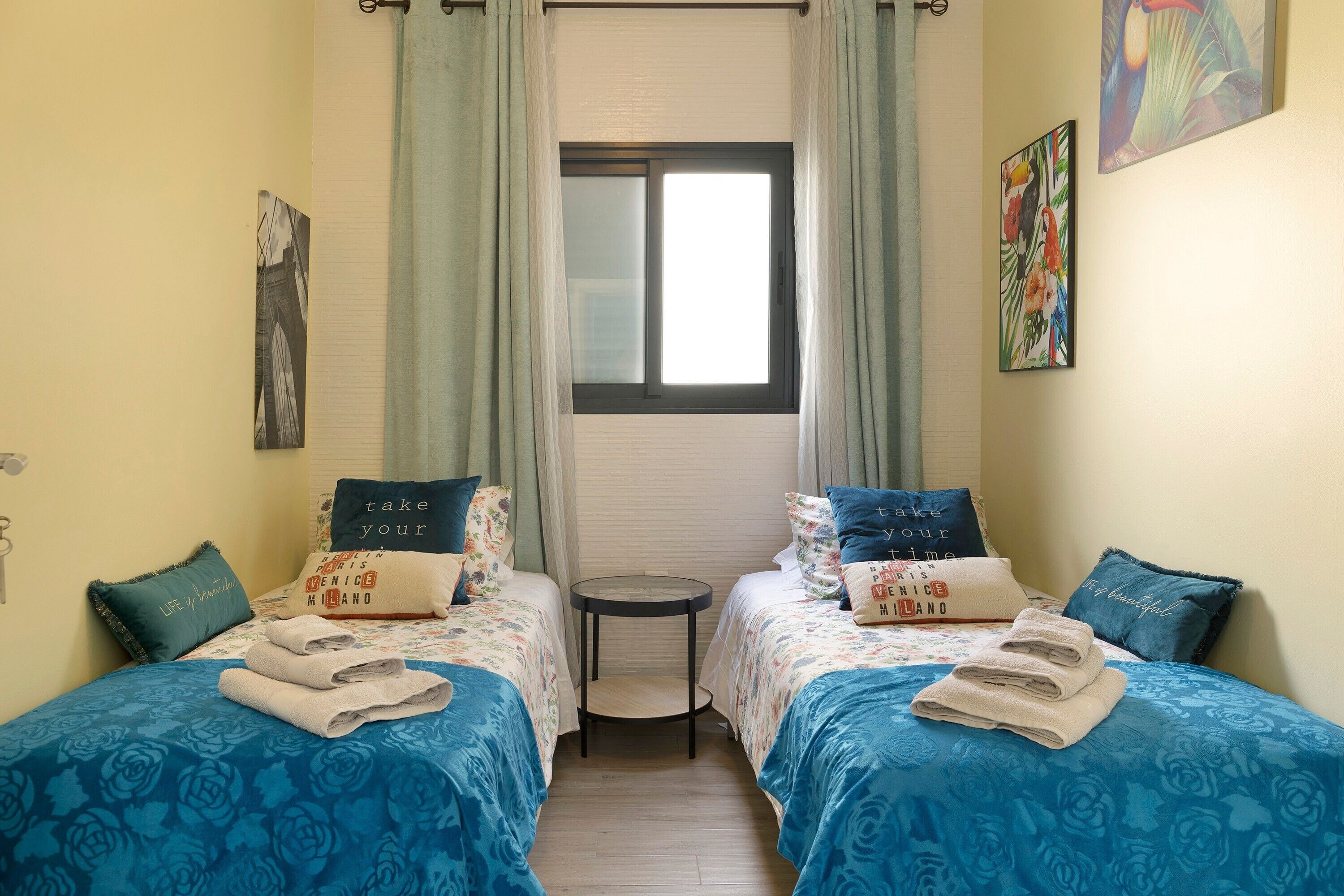 Ein Zimmer mit zwei Betten, einem kleinen Tisch, einem Fenster mit Vorhängen und Bildern an den Wänden.