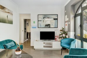 Een moderne woonkamer met een flatscreen televisie, turquoise stoelen, een witte tv-kast en een schilderij aan de muur.