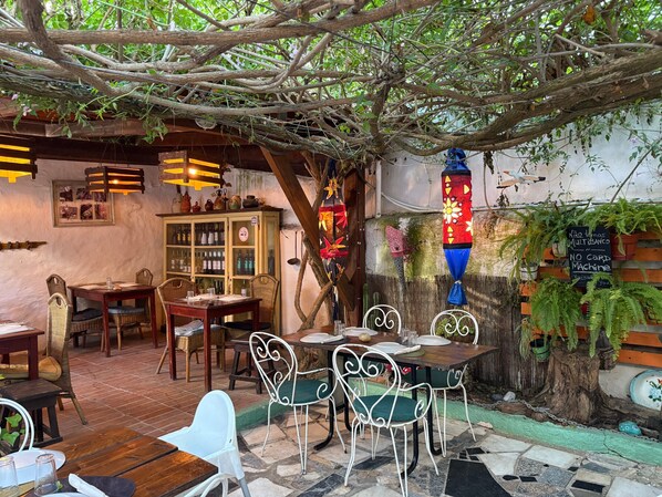 An outdoor café with tables and chairs, a wooden cabinet with bottles, and a sign indicating a no-cap machine.