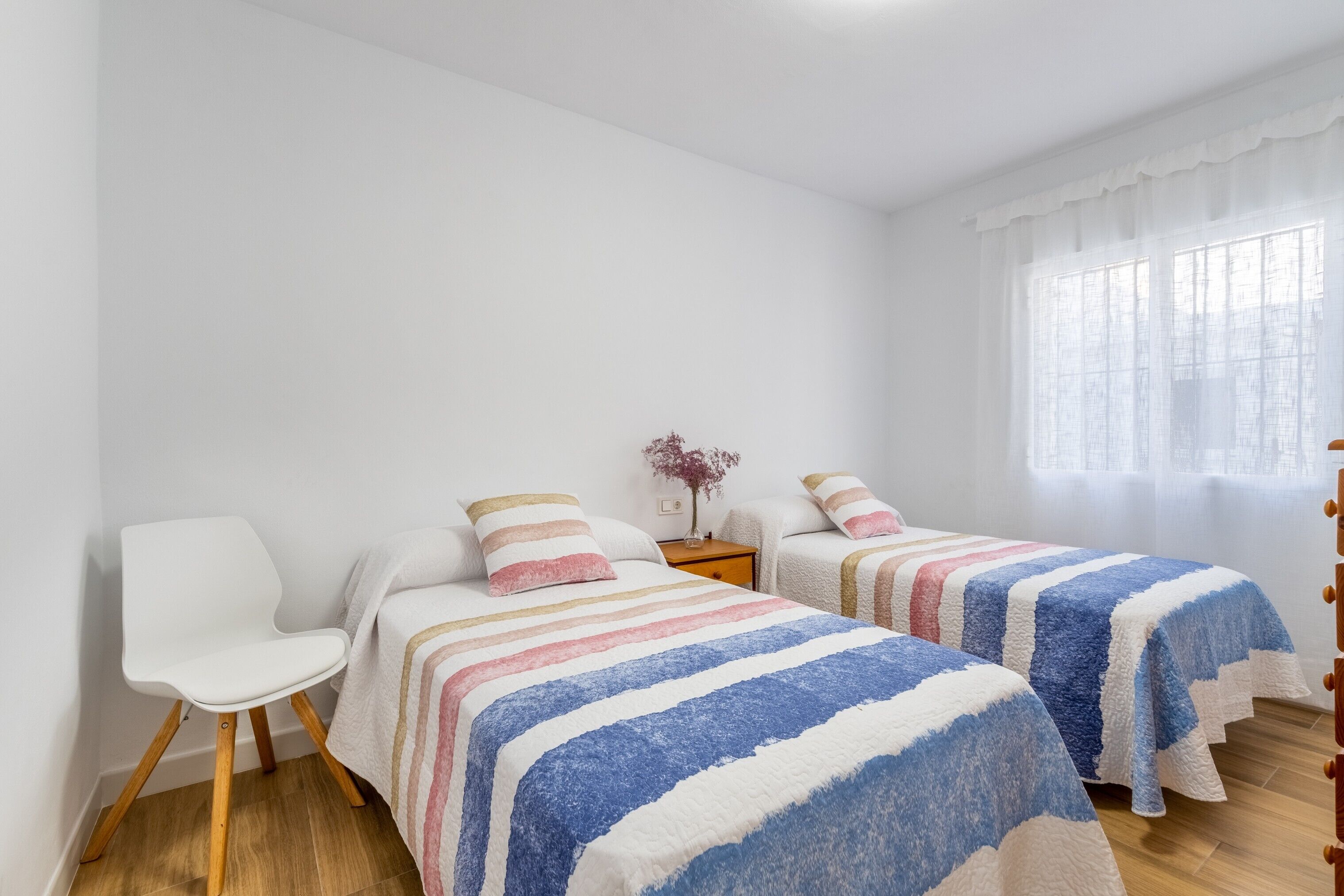 Ein Zimmer mit zwei Betten, einem Sessel, einem Nachttisch und einem Fenster mit durchsichtigen Vorhängen.