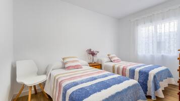 Ein Zimmer mit zwei Betten, einem Sessel, einem Nachttisch und einem Fenster mit durchsichtigen Vorhängen.
