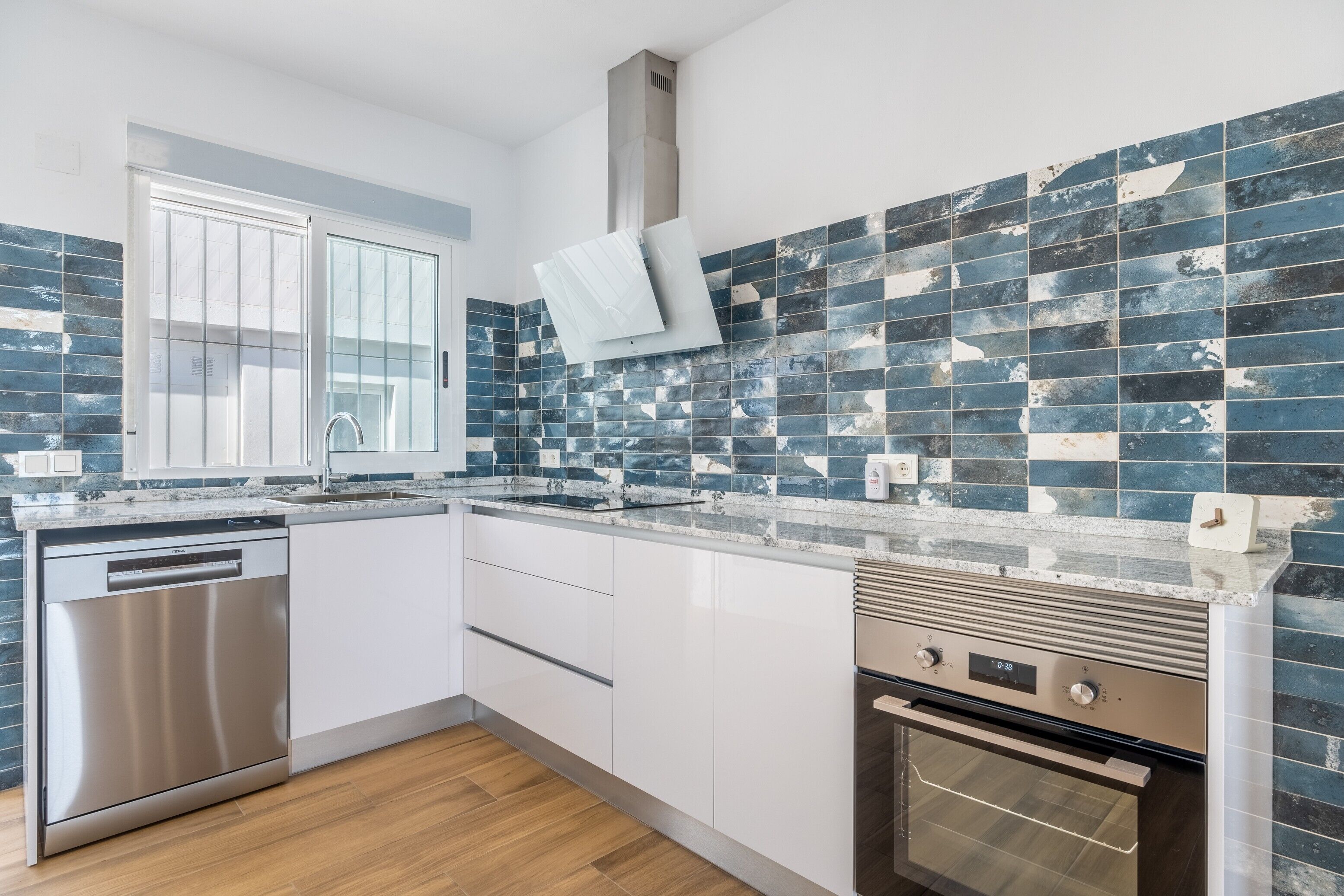 Una cocina moderna con armarios blancos, lavavajillas de acero inoxidable, horno empotrado y salpicadero con azulejos azules.