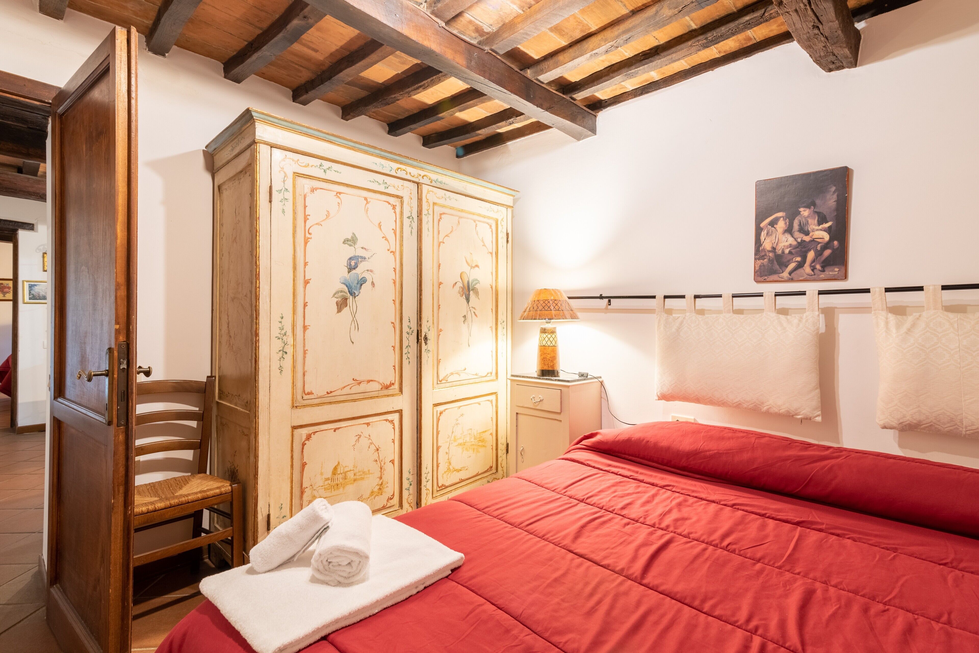Een slaapkamer met een houten garderobe, een bed met een rode sprei, een stoel, een lamp en een schilderij aan de muur.