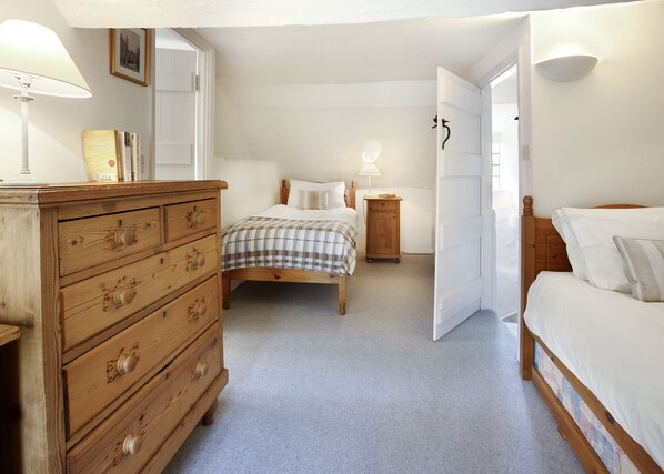 Une chambre avec une commode en bois, un lit recouvert d’une couverture à carreaux, une table de chevet avec une lampe, et une porte donnant sur une autre pièce.