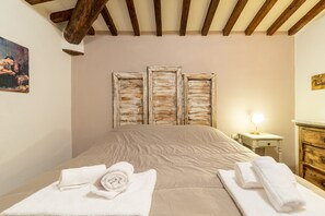 A bedroom with a bed, three wooden-framed windows, a nightstand with a lamp, and a wooden dresser.