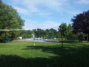 A grassy area with a volleyball net, a swimming pool, and trees.