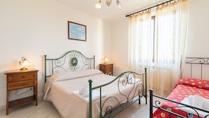 Une chambre avec un lit en métal vert, une table de chevet en bois, une petite table de nuit avec une lampe et une fenêtre avec des rideaux.