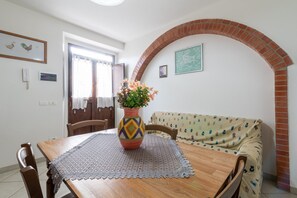 A dining area with a wooden table, a lace table runner, a colorful vase with flowers, and a patterned sofa.