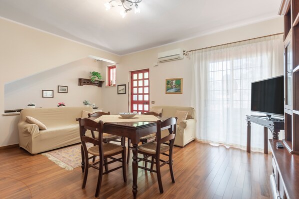 A living room with a dining table, two chairs, a sofa, a TV stand, and a window with curtains.