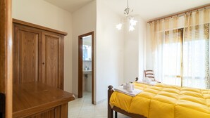 Une chambre avec une armoire en bois, un lit avec une literie jaune, un lustre et une salle de bain visible à travers une porte ouverte.