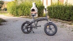 Un vélo électrique à assistance au pédalage, immobile, est garé sur un chemin de gravier, avec un four en pierre en arrière-plan.
