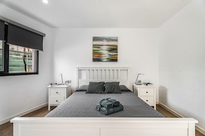 Une chambre à coucher avec un lit, deux tables de chevet, une fenêtre avec un store, un tableau au mur et une vue sur l’extérieur.