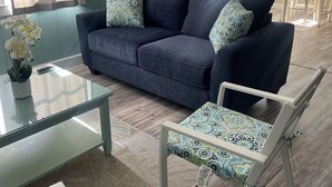Una sala de estar con un sofá azul, cojines estampados, una mesa de centro de vidrio y una silla blanca con un cojín estampado.