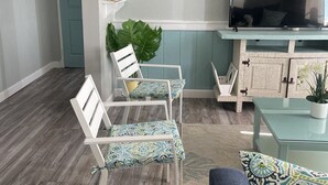 Una sala de estar con televisor, sillas blancas con cojines estampados, un sofá azul y un mueble de madera.
