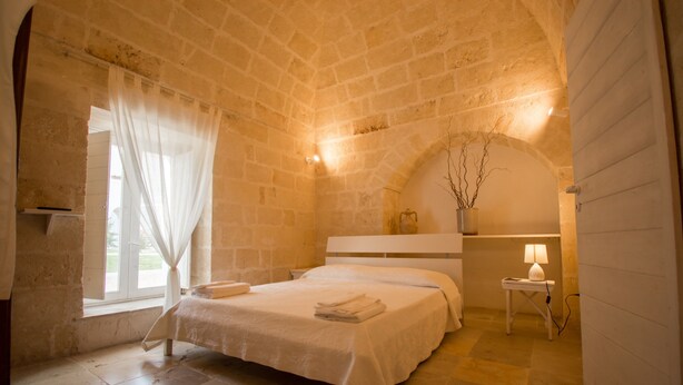 Un dormitorio con una cama, una mesita de noche, una lámpara, una ventana con cortinas y una pared de piedra.