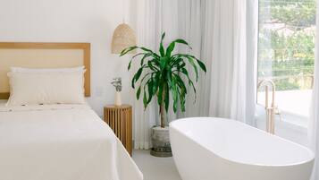 Een moderne slaapkamer met een groot bed, een vrijstaand bad, een kamerplant en een groot raam met gordijnen.