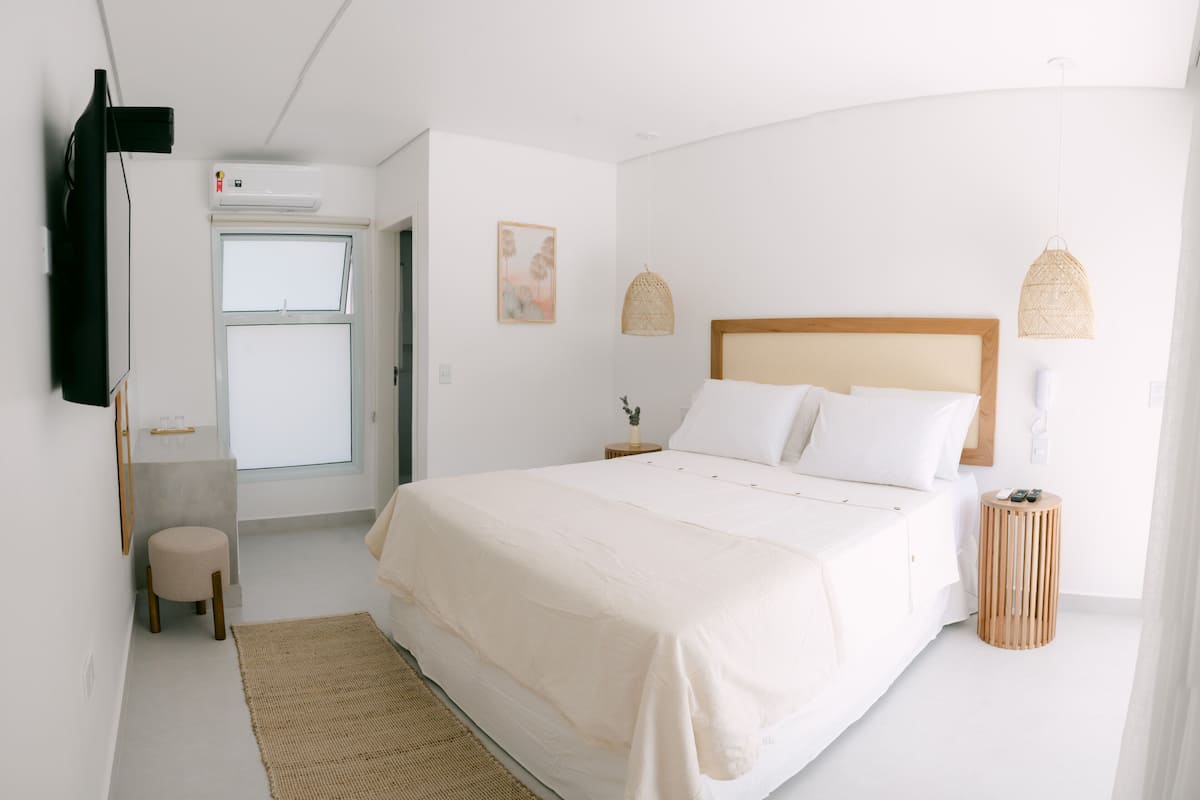 A minimalist bedroom with a large bed, bedside tables, a small stool, a wall-mounted TV, and a door leading to a bathroom.