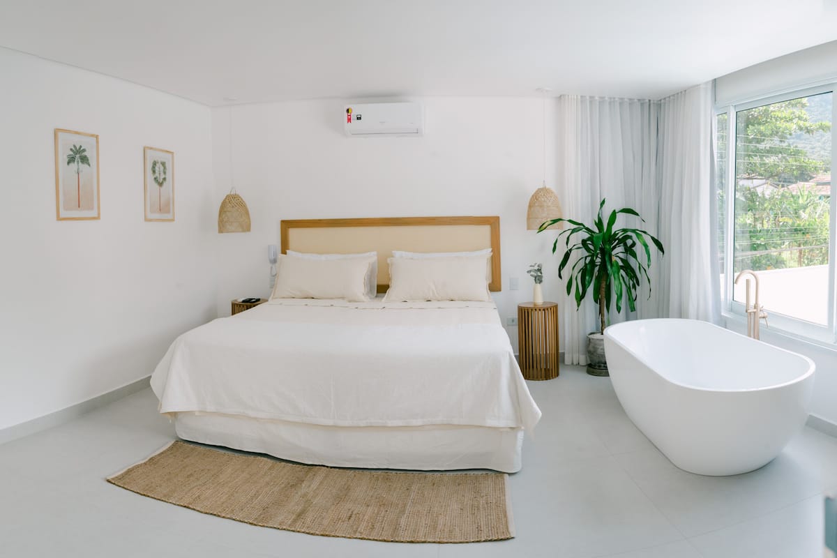 A modern bedroom with a large bed, a freestanding bathtub, and a large window with a view of greenery.
