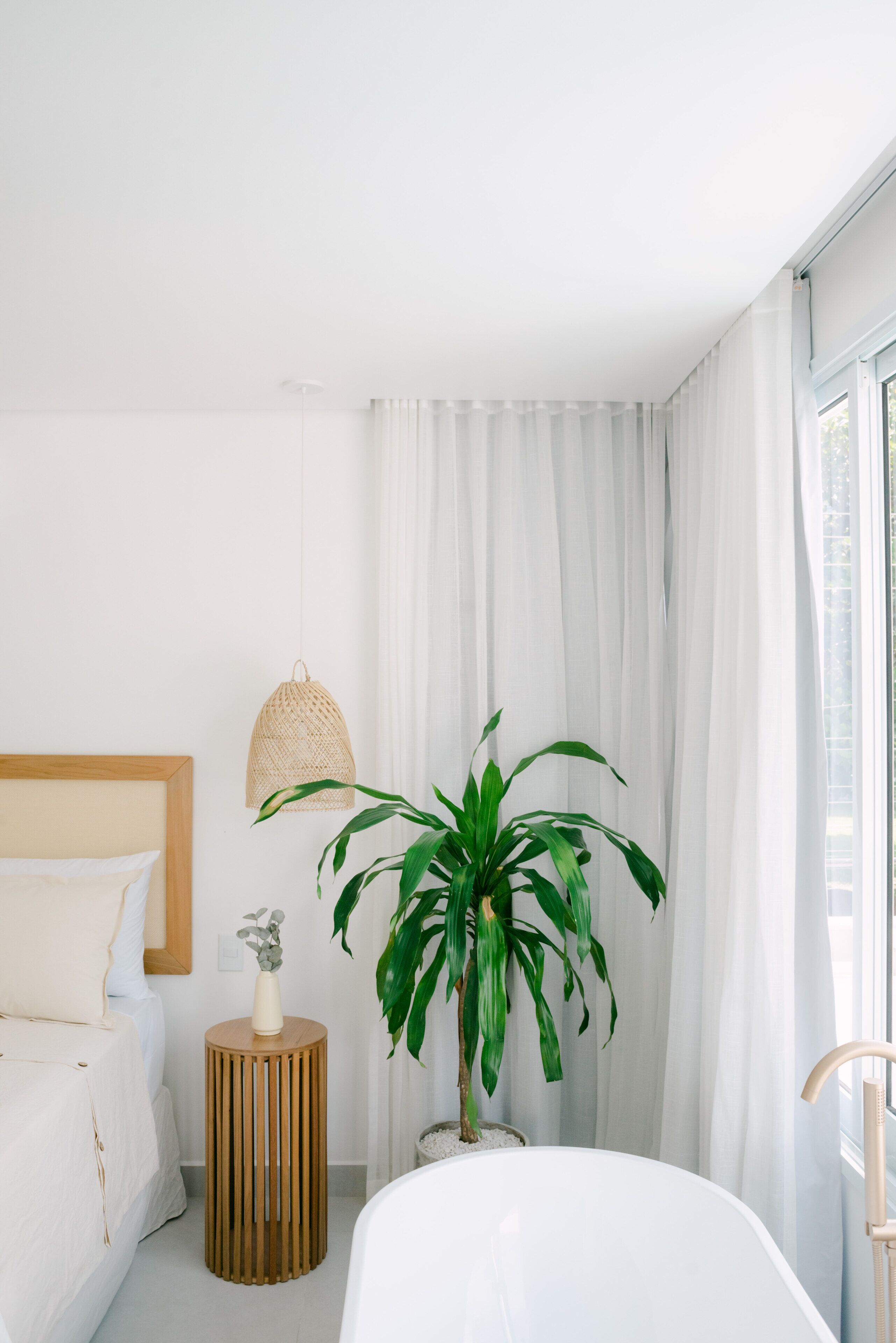 A modern hotel room with a bed, a round wooden side table, a potted plant, and a large window with curtains.