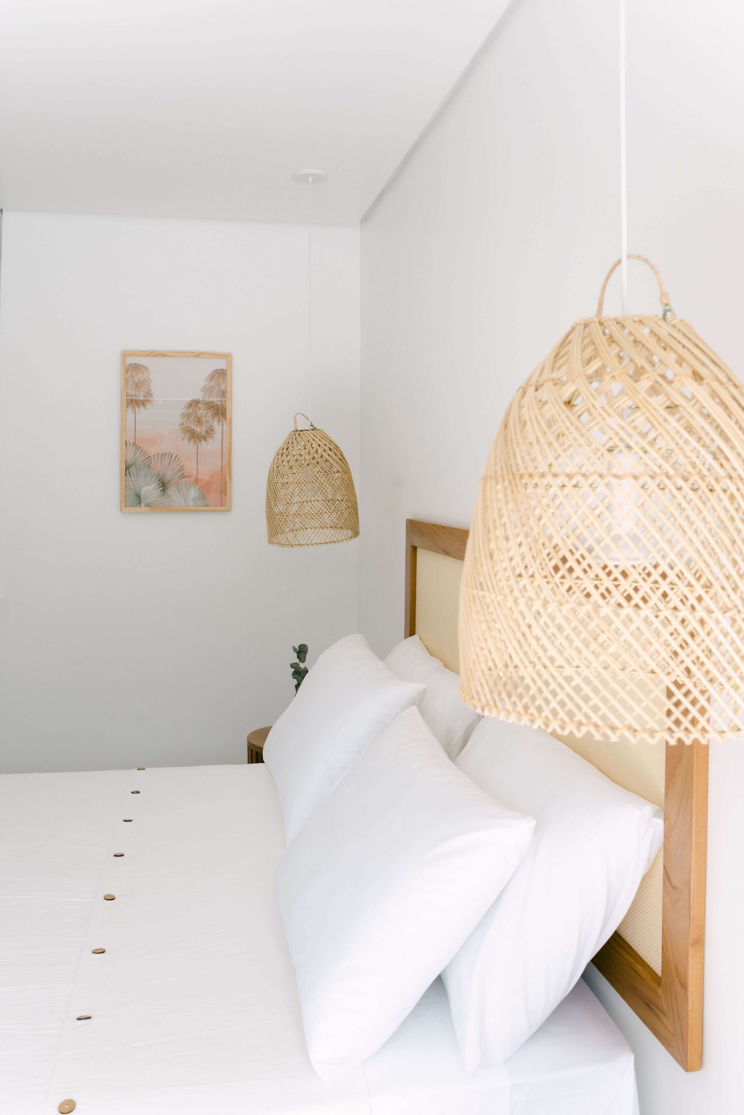 Una cama con ropa blanca y un cabecero de madera, una lámpara colgante tejida y un cuadro en la pared.
