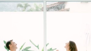 Um casal sentado em uma mesa redonda branca, com vista para uma piscina e vegetação lá fora.