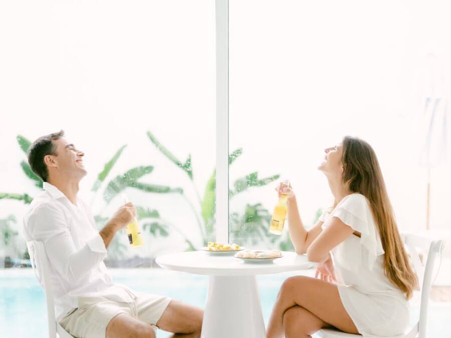 Una pareja sentada en una mesa redonda blanca, con vista a una piscina y follaje exterior.