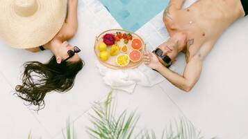 Duas pessoas deitadas sobre uma toalha branca junto a uma piscina, com uma cesta de frutas no meio.