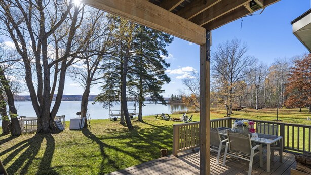 A wooden deck with outdoor furniture overlooking a lake, surrounded by trees and a grassy area. - The Sweet Seclusion Cottage with 100 feet of Private frontage Pickerel Lake! (Alanson)