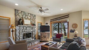A living room with a stone fireplace, a television, and a large window with a view of trees. - The Sweet Seclusion Cottage with 100 feet of Private frontage Pickerel Lake! (Alanson)