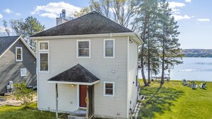 A two-story house with a front porch, a red door, and a view of a lake. - The Sweet Seclusion Cottage with 100 feet of Private frontage Pickerel Lake! (Alanson)