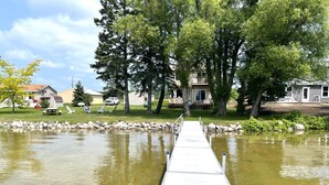 A wooden dock extending into a calm body of water, surrounded by trees and residential buildings. - The Sweet Seclusion Cottage with 100 feet of Private frontage Pickerel Lake! (Alanson)
