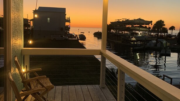 Una terraza de madera con sillas, barandilla y luces colgantes contra un fondo de atardecer.