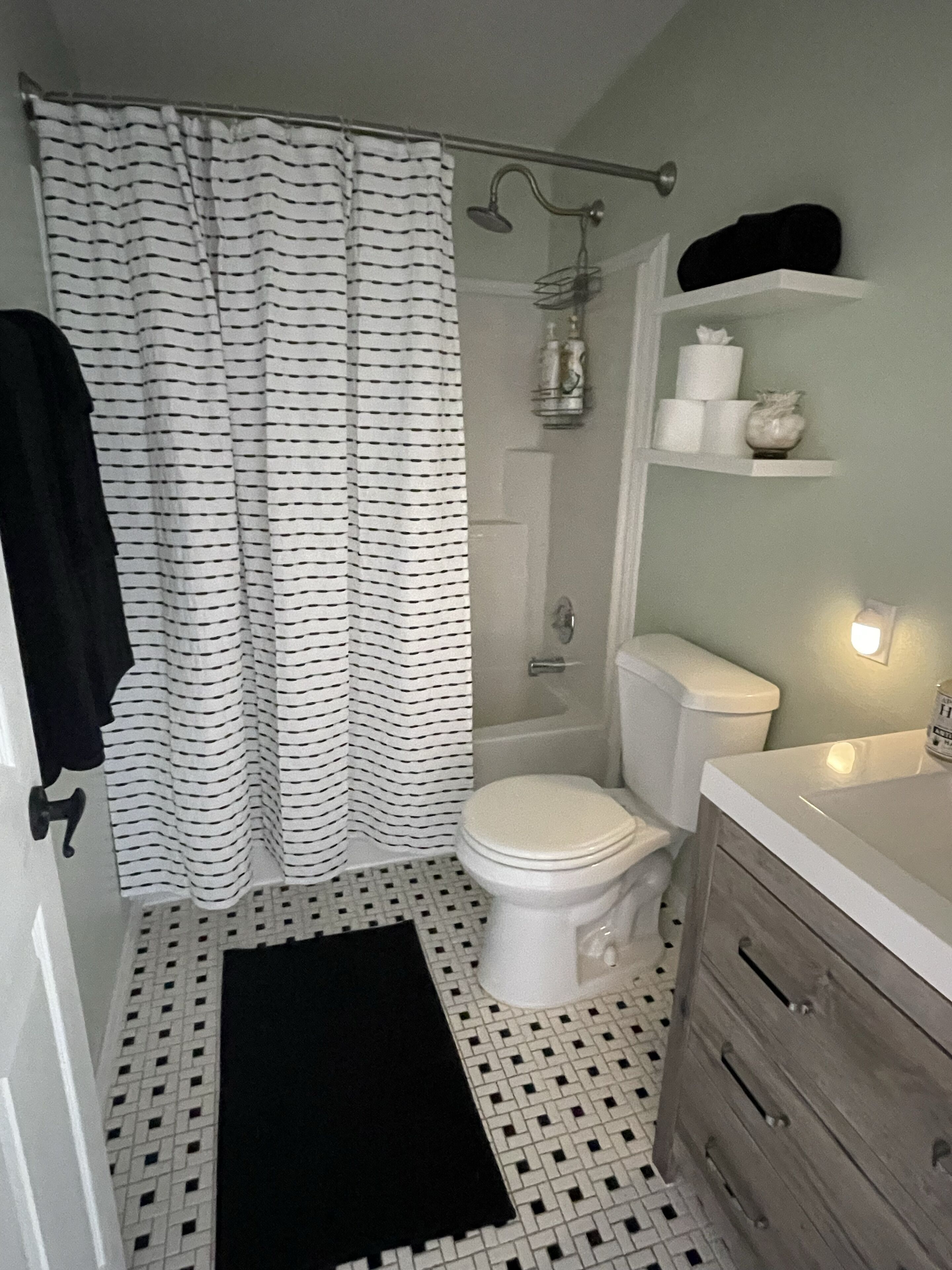 Et badeværelse med et hvidt toilet, en bruser, en badeforhæng i sort og hvid strib, en hvid vask med spejl og et hvidt håndklædestativ.