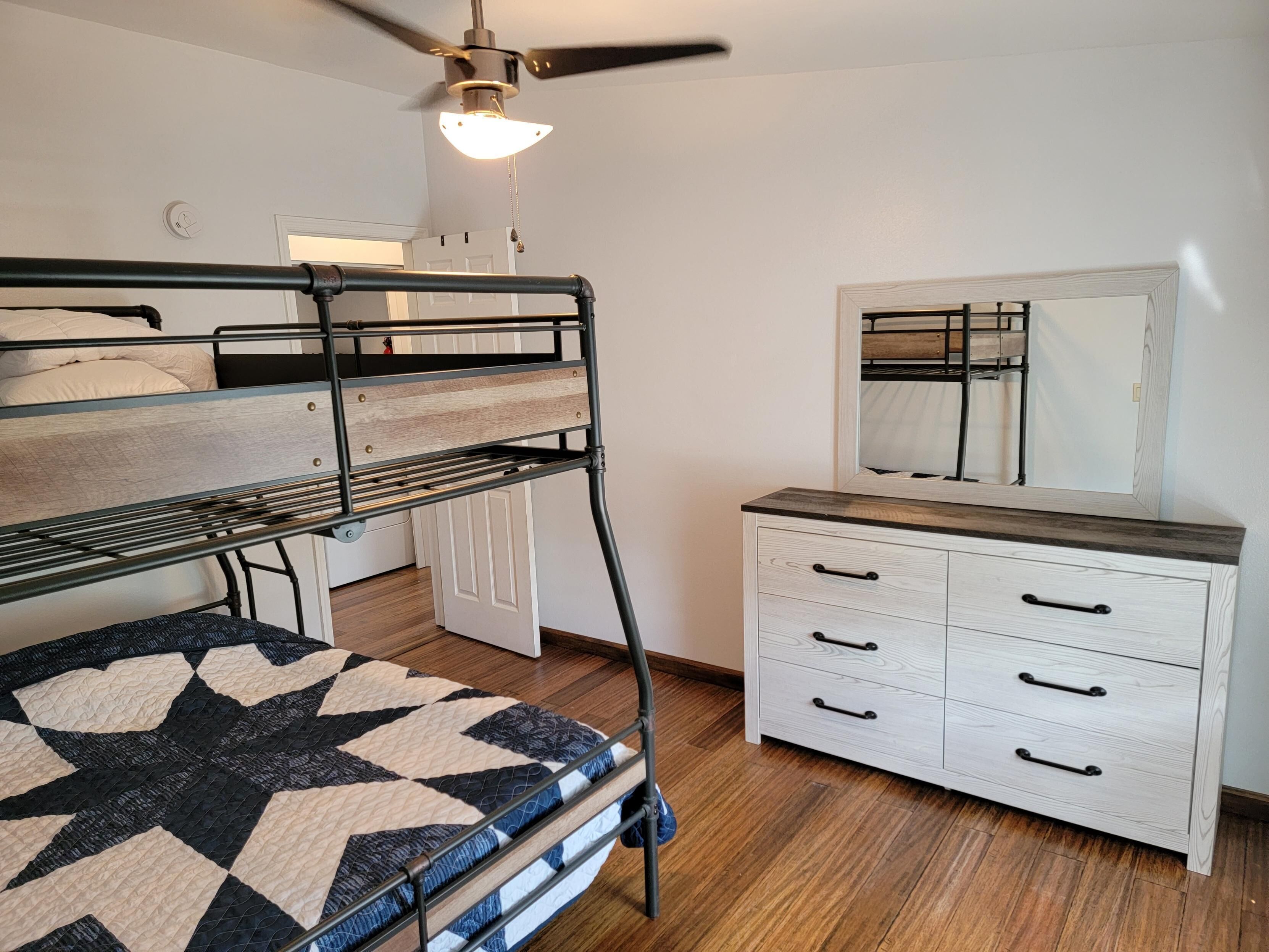 Un lit superposé avec une courtepointe à motifs géométriques, une commode blanche avec de nombreux tiroirs et un ventilateur de plafond.