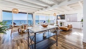 A spacious living area with a large window overlooking the sea, a dining table with chairs, a sofa, and a TV mounted on the wall. - Casa Penelope by Oraklus (Malibu)