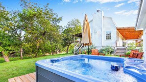 Un jacuzzi dans un jardin, avec une terrasse en bois, un patio couvert et une maison blanche en arrière-plan.