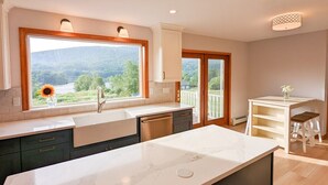 Une cuisine moderne dotée d’un grand îlot central, d’une fenêtre offrant une vue sur un lac et des montagnes, et d’une terrasse en bois à l’extérieur.