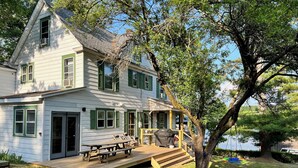 A two-story house with a porch, a wooden deck, and a large tree in the yard.