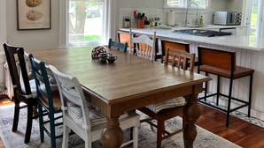 A dining area with a wooden table and chairs, a chandelier, and a kitchen in the background.