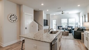 A modern kitchen with a granite countertop, stainless steel sink, and a built-in dishwasher. There is a staircase leading to another level, a dining area with a dark-colored sofa and a lamp, and a large window with blinds. - Discover why Serenity is a popular family vacation spot in Perdido Key! (Perdido Key)