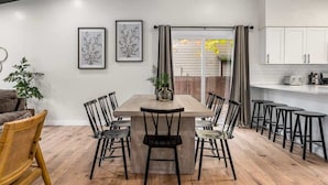 Un coin repas avec une table en bois et des chaises noires, une cuisine avec des meubles blancs et une vue sur une terrasse en bois accessible par une porte coulissante.