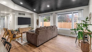 Un salon spacieux avec un plafond sombre, une grande fenêtre, un téléviseur à écran plat, un canapé marron, des chaises en bois et une plante en pot.
