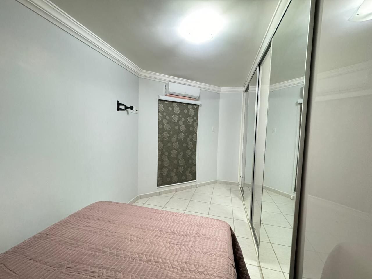 Un dormitorio con cama, armario, ventana con cortinas estampadas y equipo de aire acondicionado.
