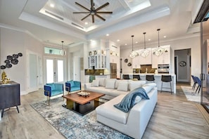 A spacious living room with a large sofa, a coffee table, and a dining area with chairs. There is a kitchen in the background with modern appliances and pendant lights. - Luxury canal home w/ heated pool & spa! (Marco Island)