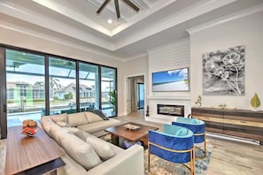 A spacious living room with a large sofa, a coffee table, and a fireplace. There are large windows offering a view of the outside. - Luxury canal home w/ heated pool & spa! (Marco Island)