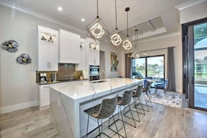 A modern kitchen with a central island, bar stools, pendant lighting, and a view to the outside. - Luxury canal home w/ heated pool & spa! (Marco Island)